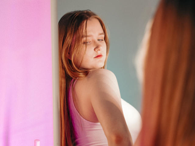 BonnieBell with long red hair, pink tank top, looking over her shoulder, showing her ass, reflection in mirror, soft light, slight blush on her cheeks, gentle expression.