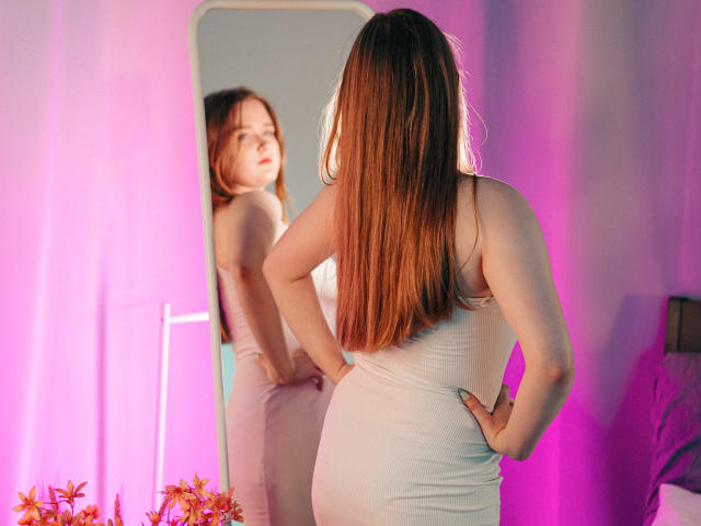 BonnieBell with long brown hair, facing mirror, wearing tight white dress, showing her ass, looking at reflection, pink and purple lighting, hands on hips, flowers in bottom left.