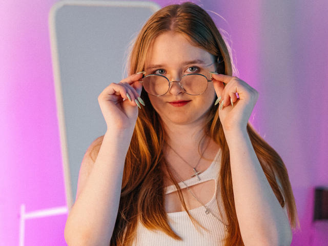 BonnieBell with long red hair, holding round glasses, wearing a white ribbed tank top, and a cross necklace, looking into the camera with a slight smile.