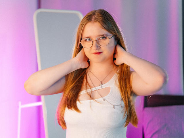 BonnieBell with long red hair, glasses, white ribbed tank top, hands on ears, necklace, pink and purple lighting, standing in front of a white frame.