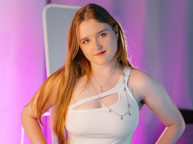 BonnieBell has long red hair, white tank top with cut-out, showing her tits, necklace with cross and small chains, pink and purple background, hands on hips, slight smile.