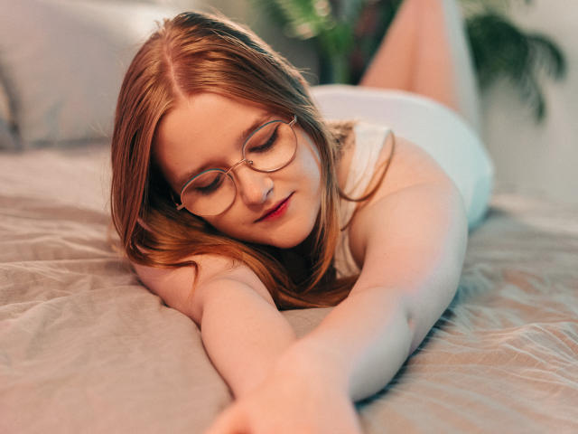 BonnieBell, a red-haired chick with glasses, lies on a bed, stretching out with her tits down and ass up, wearing white underwear, looking relaxed and cute.