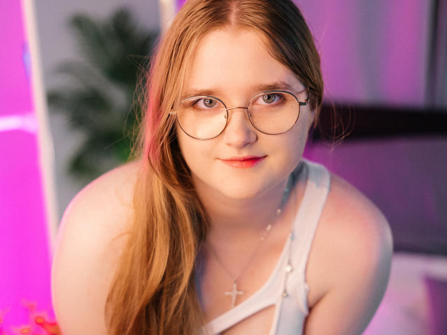 BonnieBell with long red hair, glasses, and a white strap top, showing her tits and wearing a cross necklace, looking at the camera with a slight smile.