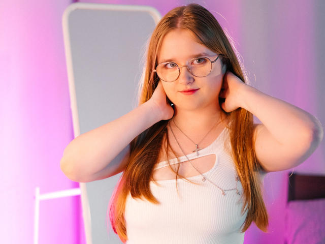 BonnieBell, a fair-skinned redhead with glasses, touches her ears, wearing a white ribbed tank top with a cutout, standing in a room with pink and purple lighting.