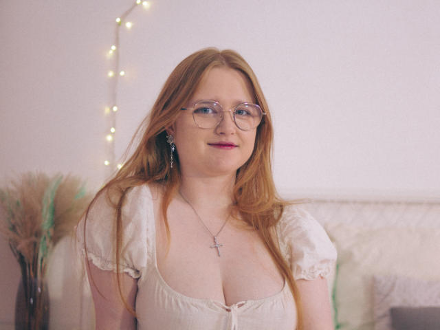 BonnieBell with red hair, glasses, and a cross necklace, smiling, wearing a white, low-cut, puffed-sleeve top, showing her tits. Soft lighting, cozy setting.