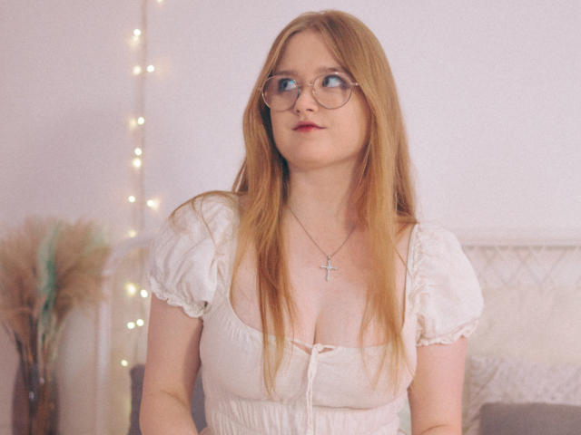 BonnieBell with long red hair, glasses, and a white dress with puffy sleeves, showing her tits, wearing a cross necklace, looking to the side.