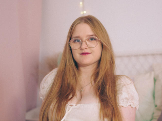 BonnieBell with long red hair, glasses, white dress, and pink lipstick, sitting, looking at camera, slight smile, cross necklace, soft light, gentle expression.