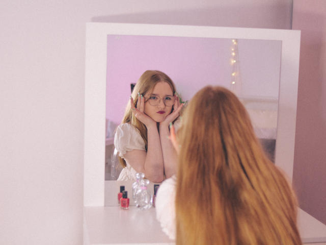 BonnieBell with long red hair, glasses, and white dress, stares at mirror, touching cheeks, showing small tits, reflection of face, white dresser with bottles, soft pink room.