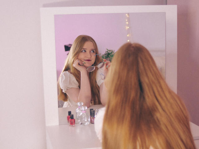 BonnieBell with long red hair, wearing a white top, looking in a mirror, holding glasses, smiling, with makeup and beauty products on a white dresser in front.