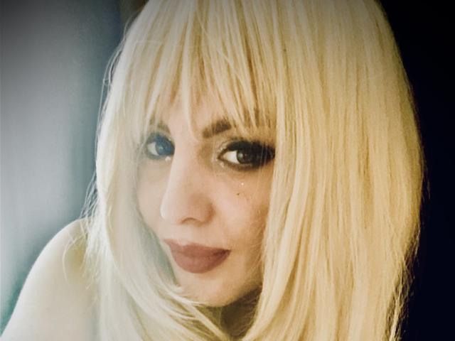 Blonde TheaPriscilla with shiny hair, dark lipstick, and slightly upturned nose, looking at the camera with a subtle, almost shy smile. Her eyes are slightly teary.