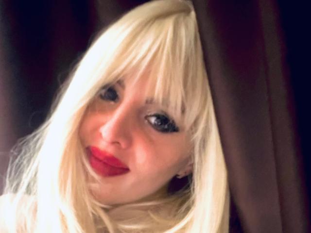 TheaPriscilla heeft blond haar, rode lippenstift en blauwe ogen, en kijkt met een lichte glimlach door donkerbruine gordijnen. Haar borsten zijn gedeeltelijk zichtbaar. Ze ziet er schattig uit..