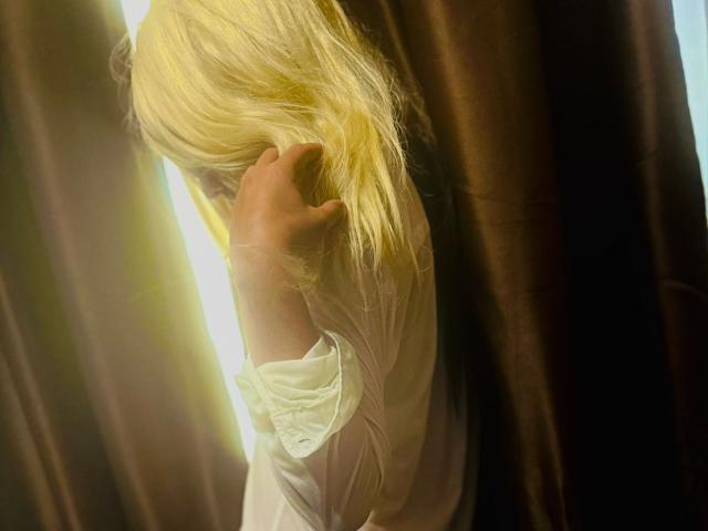 Thea Priscilla, avec ses cheveux blonds qui lui cachent les yeux, porte une chemise blanche et a une main dans ses cheveux, debout devant des rideaux marron, avec la lumière du soleil qui entre par le côté..