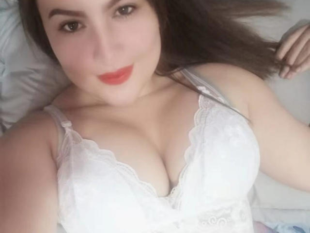 Aimi69 with pale skin, brown hair, and red lipstick, lying on a bed, wearing a white lace bra that shows her big, perky tits. Her right hand is near her head.