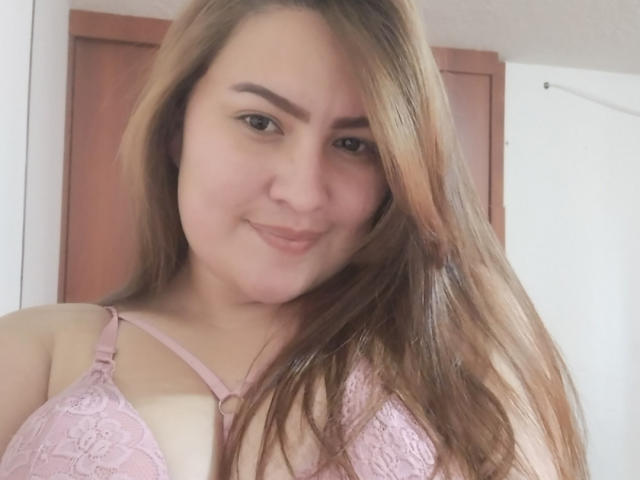 Aimi69 met lang bruin haar, lichte huid en een roze kantbeha, lacht naar de camera en laat haar borsten zien. Ze is schattig en heeft een subtiele grijns..