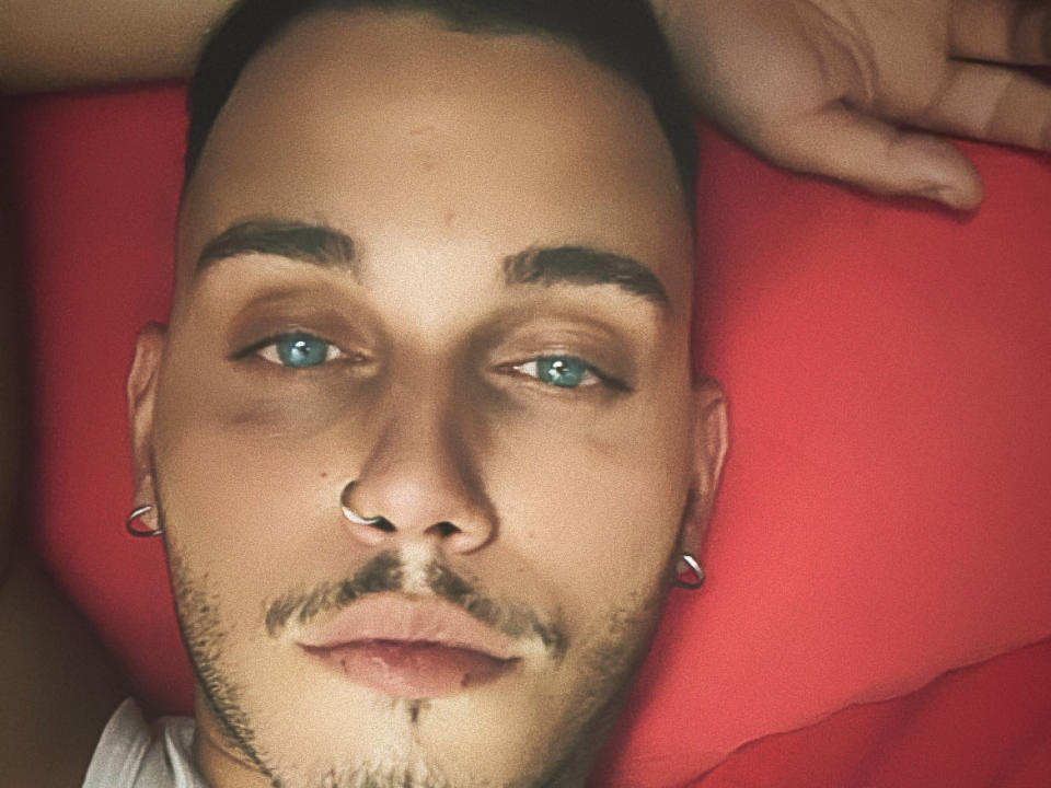 BigHotDick69 has blue eyes, a nose ring, and earrings, with a short haircut, beard, and mustache, lying on a red pillow, looking up with a hand near his head.