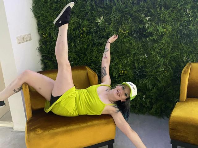 Linda-Lush-hot with pale skin, black hair, and tattoos, wears a yellow dress and white cap, lying on a yellow chair, leg up, smiling, black socks, showing her pussy.
