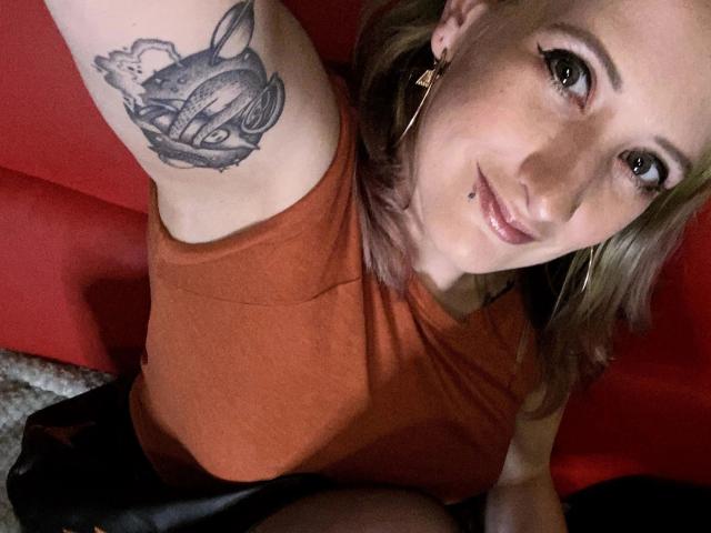 Linda-Lush-hot with a tattoo on her arm, orange shirt, black skirt, and hoop earrings, smiling at the camera, showing her nose piercing. She has light brown hair.