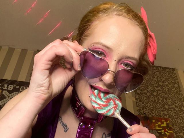 Linda-Lush-hot with pink hair, pink eyeshadow, purple heart sunglasses, pink bow, purple collar, holding a swirled pink-green lollipop, sucking on it, tattoos on chest.