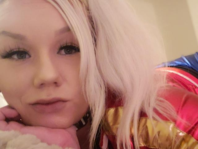 BustinBlonder with pink hair, heavy eyelashes, and pouty lips, wearing a shiny red and gold jacket, resting chin on hand, looking directly at camera.