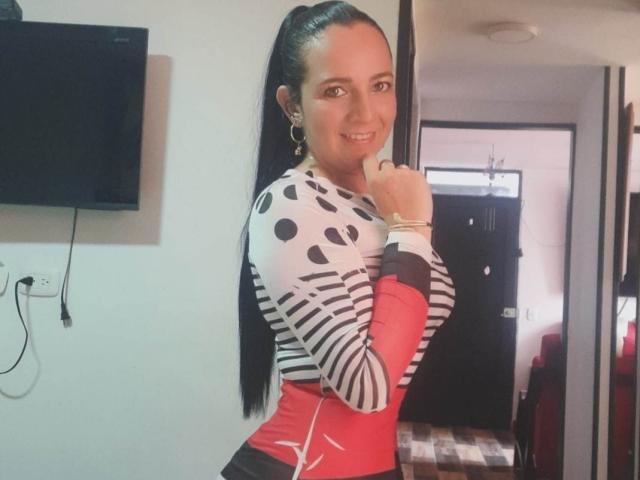 SofySexy69 with long black hair in a ponytail, wearing a white and black polka dot top with red sleeves, smiling, touching her chin, in a room with a TV.