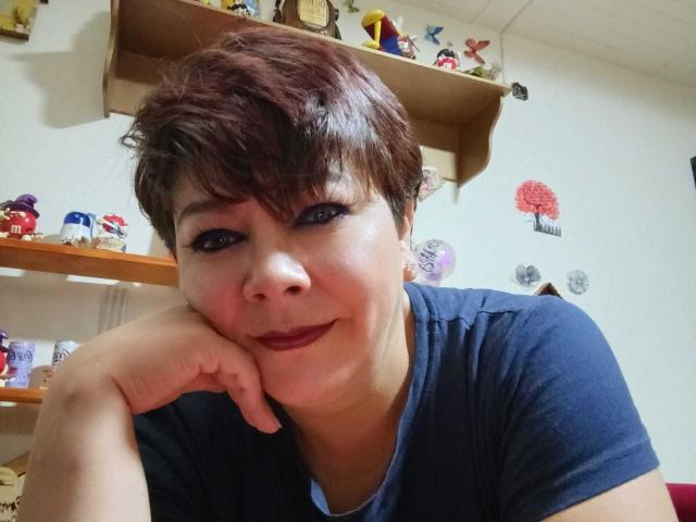 Angellovelly has short brown hair, dark blue eyes, and wears a navy shirt, resting her chin on her hand with a slight smile, showing her red lipstick.