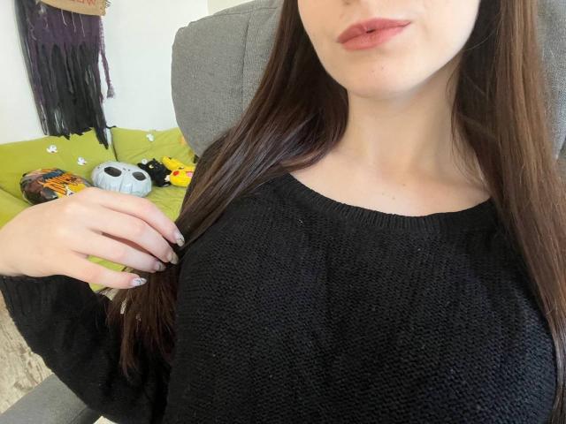 Valissia with long brown hair, wearing a black knit sweater, showing her tits, light pink lipstick, nail polish, hand on her hair, sitting in a gray chair.
