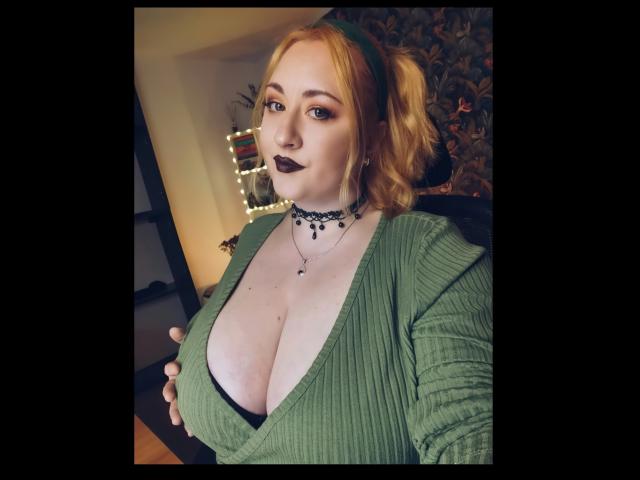 LuxNoir with big tits, green ribbed shirt, black choker, dark lipstick, and blonde puffy hair, holding her tits, looking at the camera, in a dimly lit room.