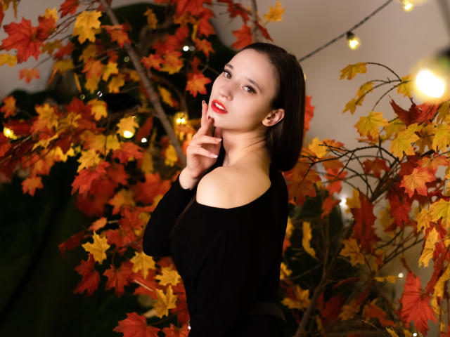 StacieLove con lápiz labial rojo, blusa negra con un hombro descubierto, cabello oscuro y con un dedo en la boca, rodeada de hojas de otoño, con luces de cadena brillantes en el fondo..