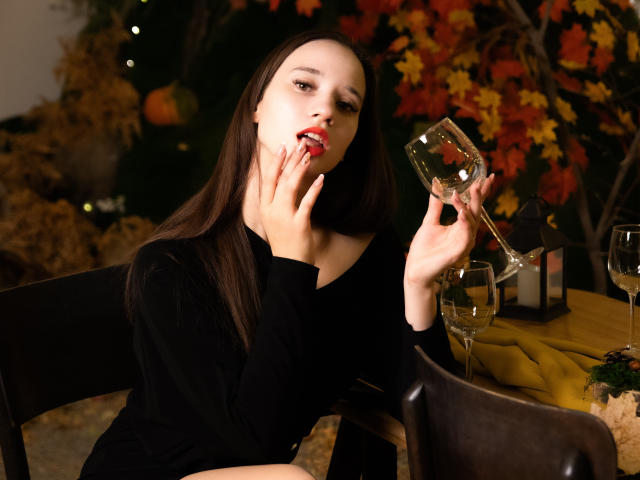 StacieLove with long brown hair, black top, holding a glass of red wine, licking her fingers, sitting at a table with a yellow cloth, wine glasses, and candles.