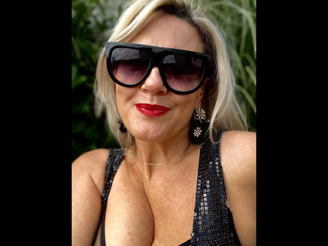 AdeleMILF-hot with big tits, wearing black sunglasses, red lipstick, and a sparkly black dress, has blonde hair and star-shaped earrings, smiling slightly.
