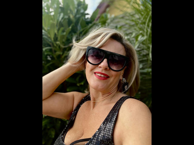 AdeleMILF-hot with short blonde hair, black sunglasses, red lipstick, and a black sequined top, showing her tits and smiling, hand in hair, surrounded by green plants.