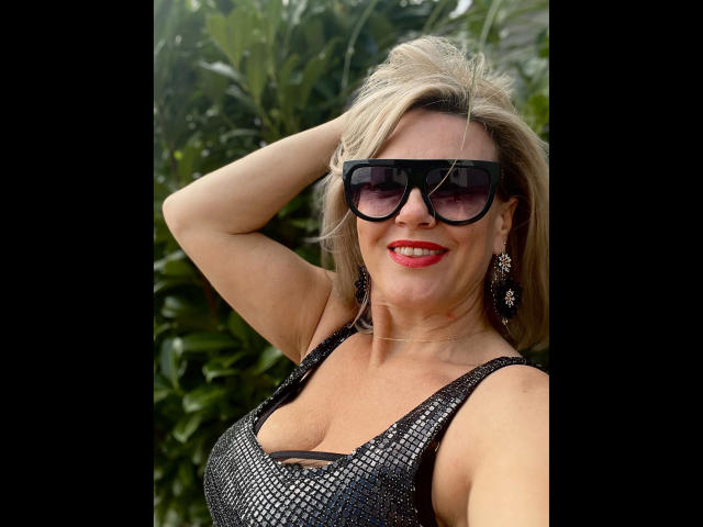 AdeleMILF-hot with blonde hair, black sunglasses, red lipstick, and a shiny black dress, showing off her tits and wearing dangly earrings, smiling confidently with one hand up.