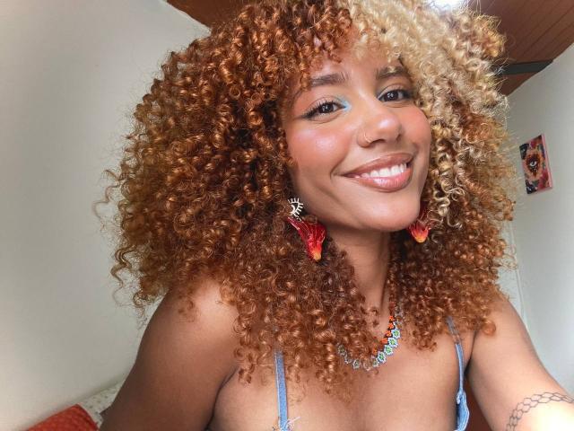 Cinthia Walker, com cabelos cacheados, loiros e castanhos, sorrindo, usando brincos de borboleta vermelhas, um colar azul e um sutiã azul, mostrando seus seios. Seu nariz é perfurado..
