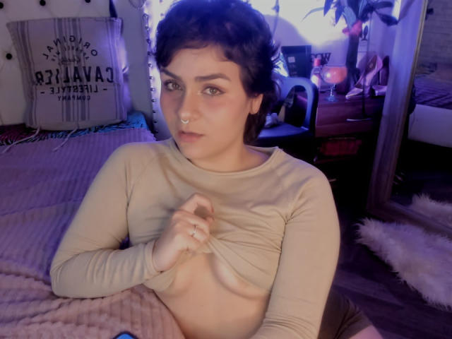 IsabelaCooper with short dark hair, pale skin, and nose ring, lifting beige long-sleeve top to expose small, perky tits, in dimly lit bedroom.
