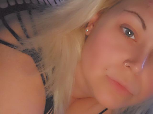 PhatKimPrincess with blonde hair, blue eyes, nose ring, small earrings, and subtle makeup, looking slightly to the side, wearing a black top, with soft, warm lighting.