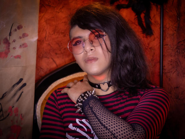 Saturni with dark wavy hair, red glasses, heart choker, striped red-black shirt, fishnet arm, spiked bracelet, looking at camera, red-lit background, medium shot, slight smirk, hands on chest.