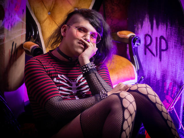 Saturni with shoulder-length dark hair, pink glasses, and a striped red-black shirt, sitting with hand on face, wearing fishnet stockings and a black choker, looking relaxed.