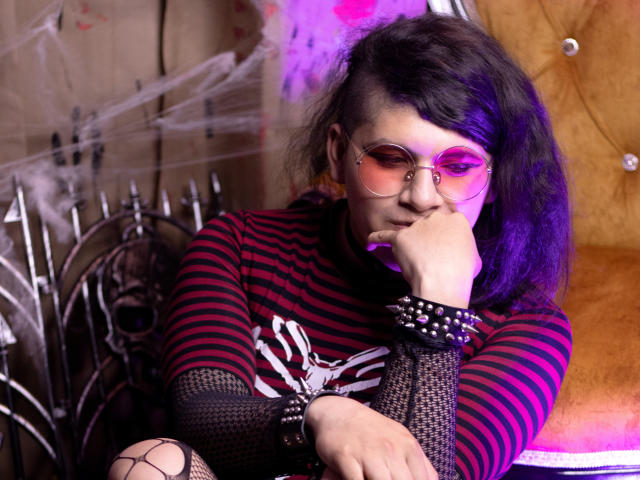 Saturni with purple-dyed hair, pink-tinted glasses, striped shirt, black fishnet sleeves, studded wristband, smoking, resting chin on hand, purple and pink lighting.