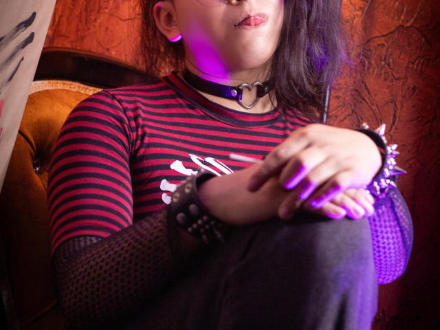 Saturni with striped red and black shirt, black choker, spiked bracelets, and purple nail polish, tongue out, hands on knees, dark hair, orange wall, and purple light.