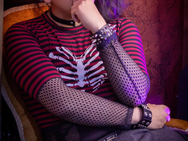 Saturni with striped red and black shirt, white bone design, fishnet sleeves, black spiked wristbands, and choker, sitting with hand near face, purple lighting.