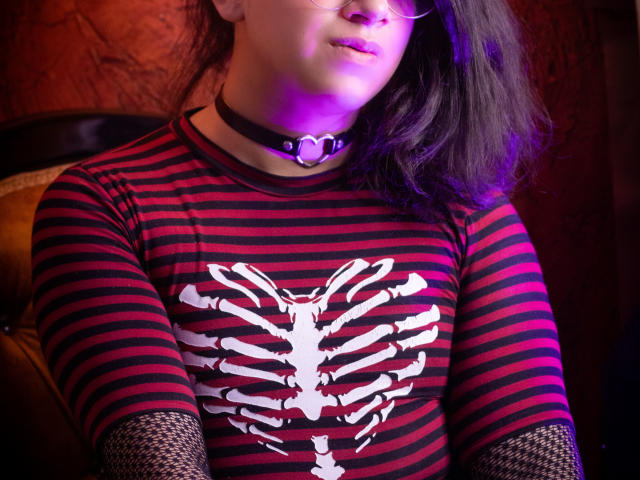Saturni with dark hair, glasses, wearing a red-black striped shirt with a white skeleton design, black choker with heart, fishnet sleeves, and purple lighting on her face.