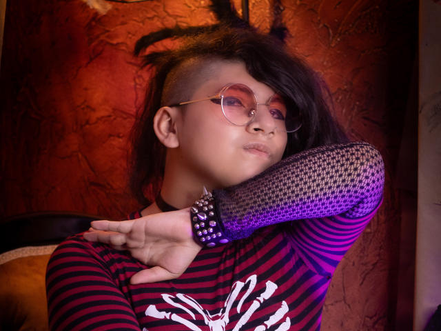 Saturni with a mohawk, wearing round glasses, striped shirt, and fishnet sleeve, showing off spiked bracelet, looking pouty. Red textured wall in background. Soft purple lighting.