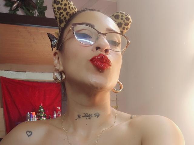LunaCandy met luipaardoren, een bril, rode lippen en een neusring, met een frons. Ze heeft oorringen, een tatoeage onder haar nek en een kleine harttatoeage op haar schouder..