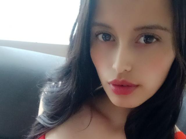 LaylaFoxy hat langes, dunkles Haar, blaue Augen, rosafarbene Lippen und helle Haut. Sie trägt ein rotes Oberteil, wobei ihre Brüste leicht sichtbar sind, und blickt mit einem leichten Lächeln in die Kamera..