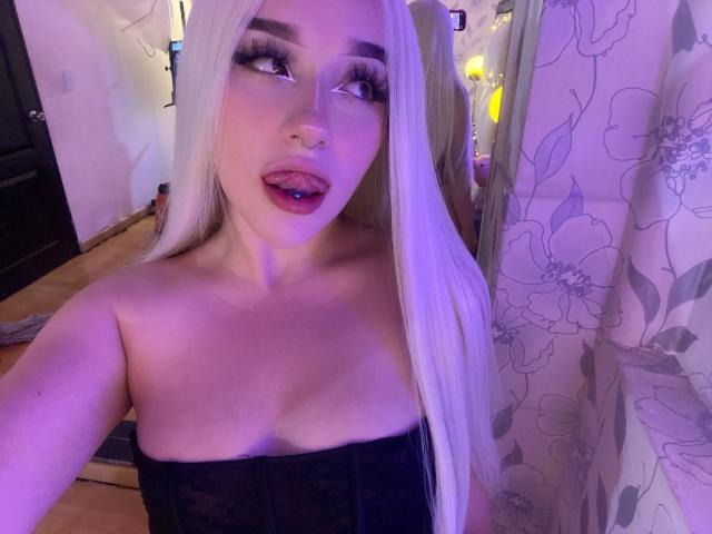 CristySanz with long blonde hair, dark makeup, purple lipstick, tongue out, black lace top, showing tits, nose piercing, in purple-lit room, floral curtain on the right.