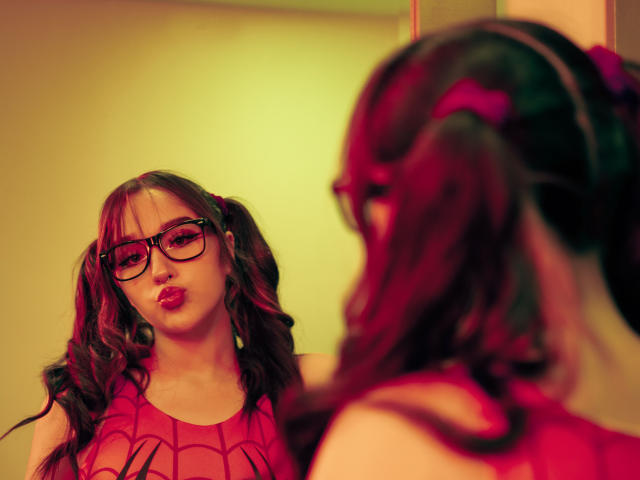 CristySanz with puffy red lips, wearing a red Spider-Man shirt, and black glasses, reflecting in a mirror, showing her curly brown pigtails and red hair ties.