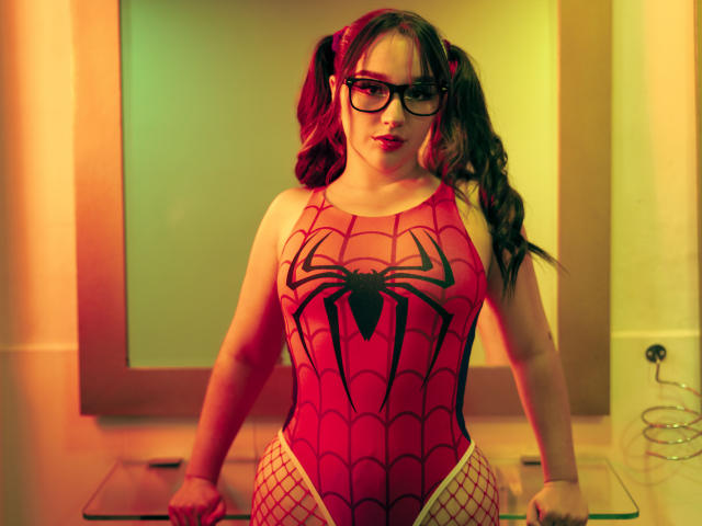 CristySanz in a red Spider-Man bodysuit with black spider logo, fishnet pants, and pigtails, wearing glasses, standing with arms on a counter, under warm lighting.