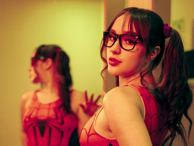 CristySanz with red spiderweb costume, pigtails, black glasses, and glossy lips, reflected in mirror, showing her back and tits, in warm, yellow-lit room.