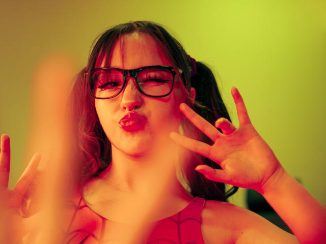 CristySanz with puffy red lips, black glasses, and pink-tinted hair in pigtails, making a "V" sign with both hands, wearing a red top, under warm green light.