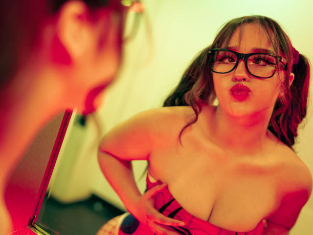 CristySanz with puffy red lips, black glasses, and brown pigtails, pulling down a red strapless top to expose her big, bare boobs in a mirror.